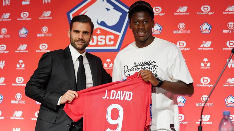 Un homme en complet tient un chandail de soccer rouge avec le joueur qui le portera avec sa nouvelle équipe. 