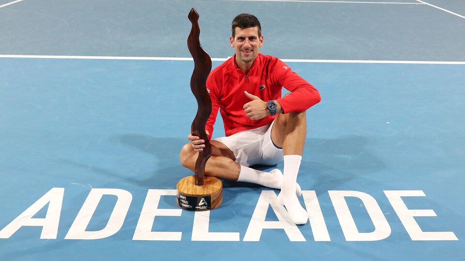 Un joueur de tennis est assis sur un terrain où il est écrit : Adelaide. Il lève un pouce en l'air et tient un trophée dans l'autre main.