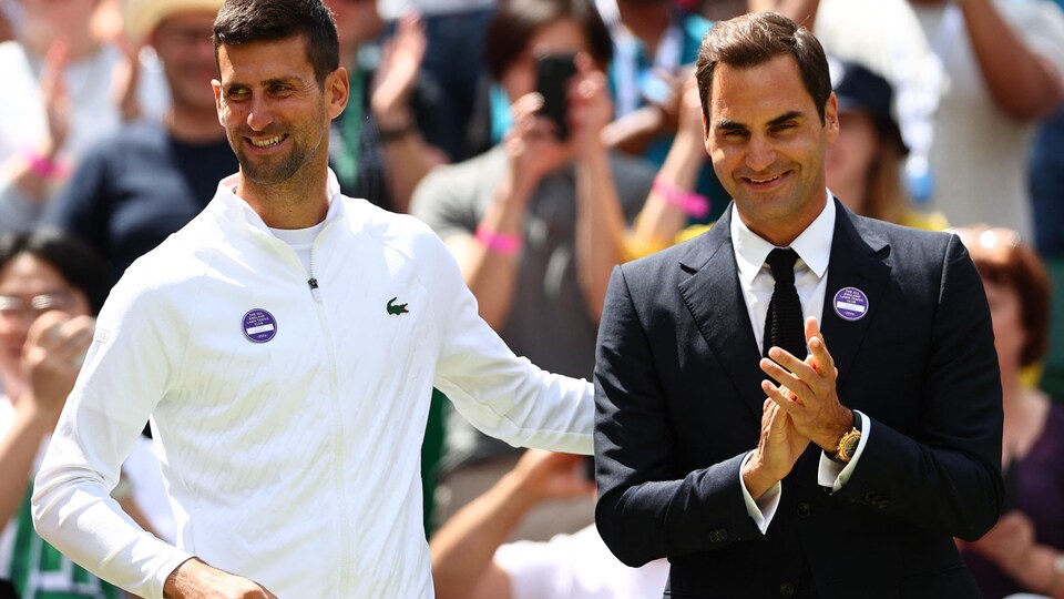 Deux joueurs de tennis sont souriants, l'un en survêtement, l'autre en costume civil. 