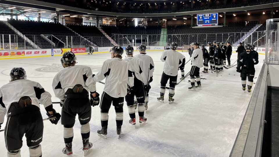 Des joueurs de hockey sont sur le côté de la patinoire et regardent deux coéquipiers participer à un exercice.