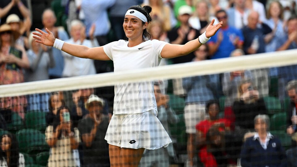 Une joueuse de tennis, en blanc, lève les bras après avoir gagné son match. Le public l'applaudit.  