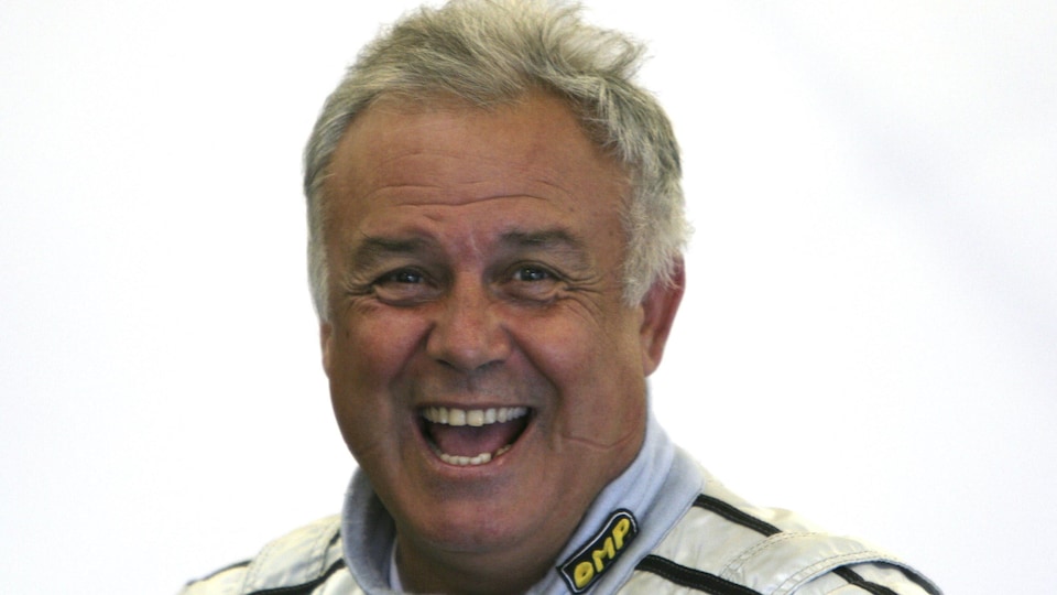 Un homme aux cheveux gris et habillé en pilote de course sourit.