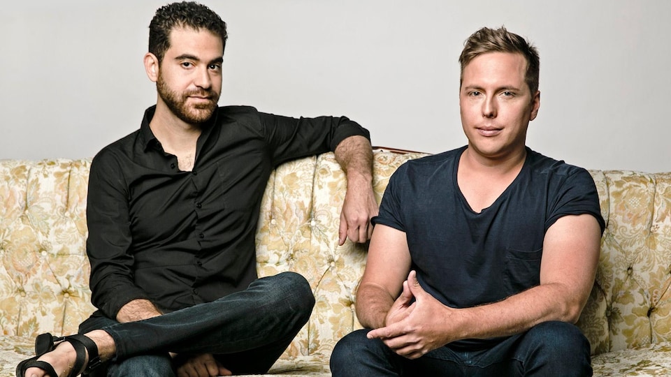 Deux hommes assis sur un divan sourient légèrement. 