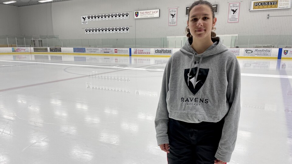 Une jeune joueuse de hockey se tient sur la patinoire, près du banc des siens et prend la pose pour la photo.