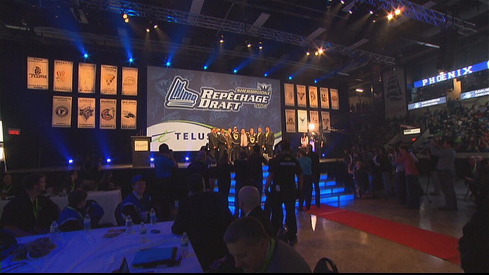 Une séance de repêchage de la LHJMQ au Palais des sports Léopold-Drolet de Sherbrooke. On y voit une équipe sur la scène devant une salle comble. 