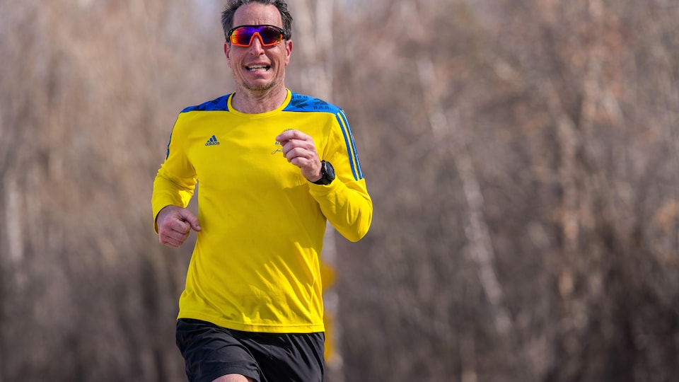 Un coureur en action près d'un petit boisé porte des lunettes de soleil et un chandail à manches longues bleu et jaune.