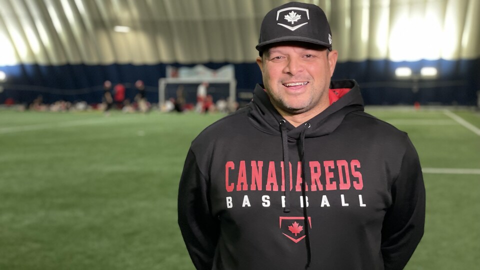 Un entraîneur de baseball habillé en noir prend la pose devant la caméra sur un terrain synthétique intérieur.