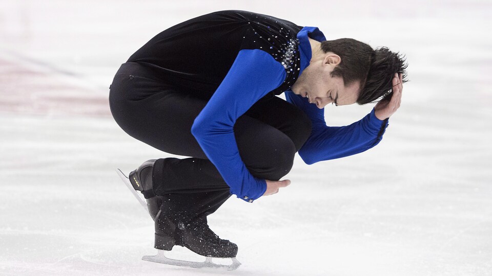Un patineur artistique a la tête penchée pendant une rotation en fin de routine. Il porte un haut noir avec des manches bleues. 