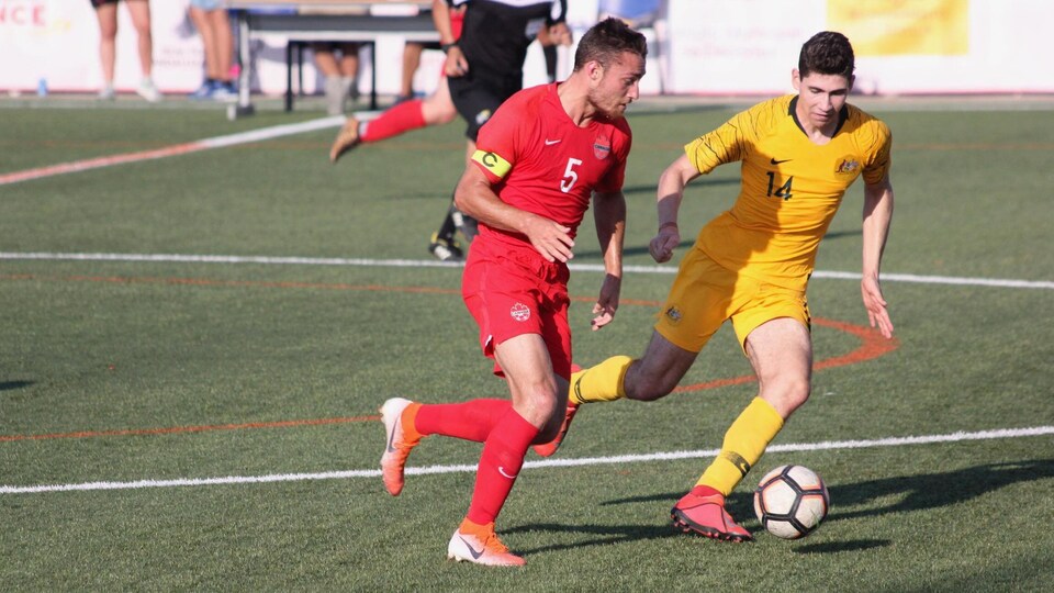 Un joueur de soccer en rouge tente de déjouer un adversaire en jaune qui le marque de près.