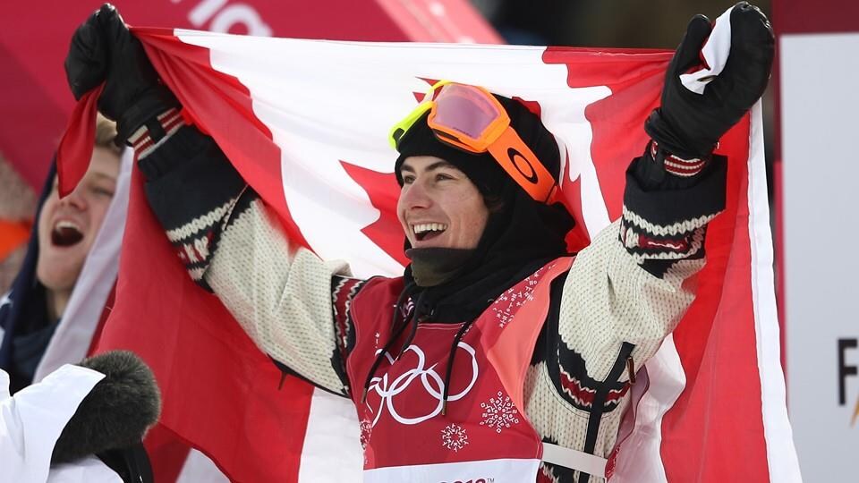 Il sourit et lève les bras en tenant un drapeau canadien sur son dos.