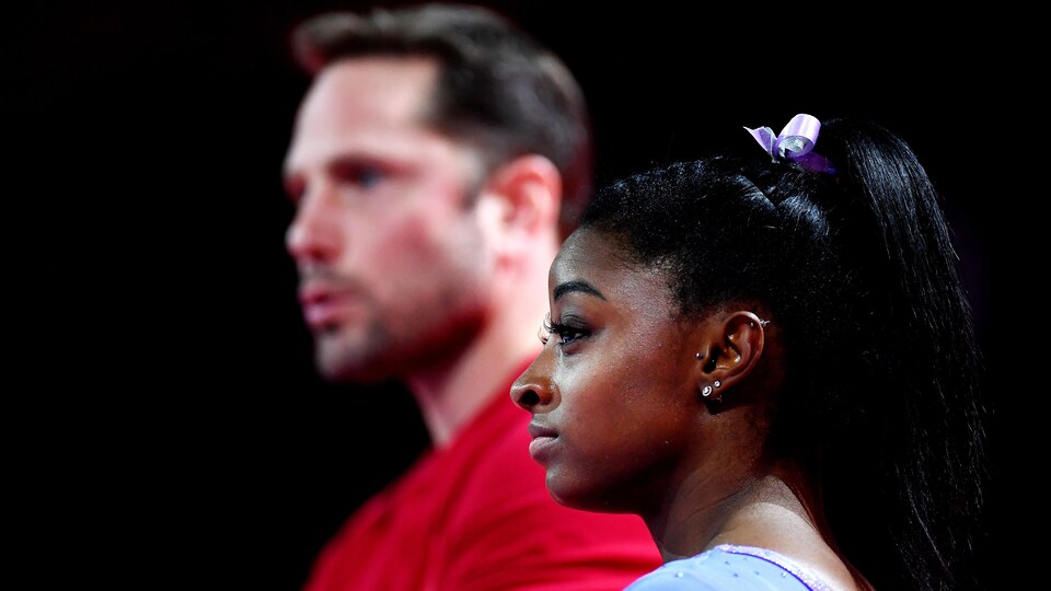 Simone Biles de profil écoute pendant qu'en arrière-plan parle son entraîneur Laurent Landi