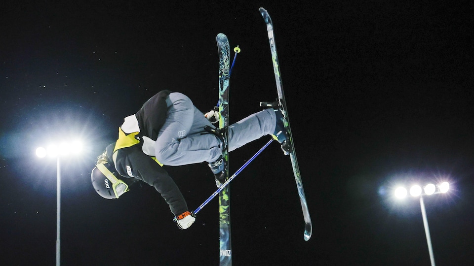 Rachael Karker effectue une figure où elle a la tête vers le sol, tout en attrapant son ski droit de la main droite.
