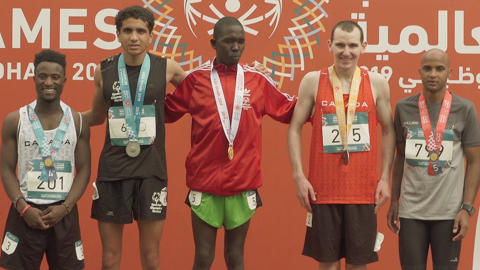 Il porte une médaille de bronze à son cou et se tient sur le podiums avec quatre autres coureurs.