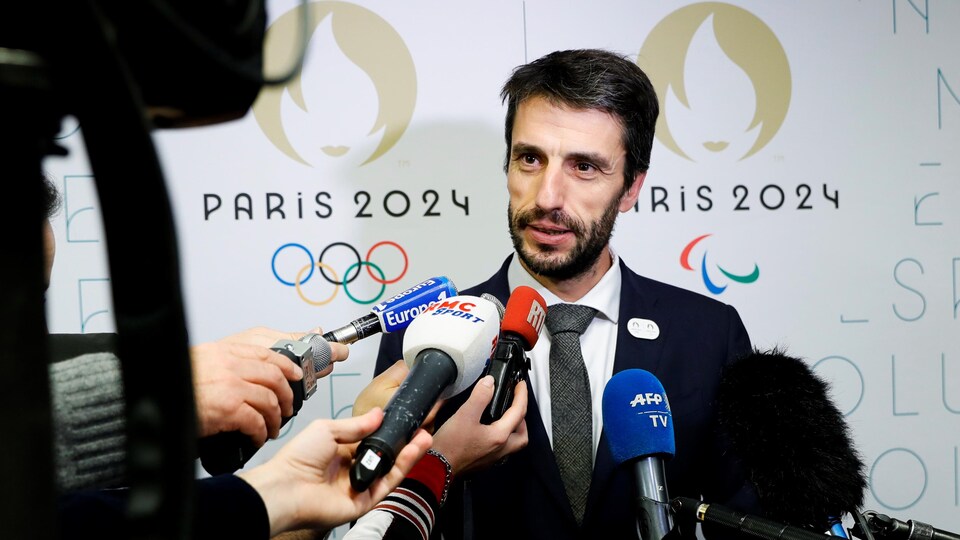 The bearded man answers questions behind numerous microphones and posters displaying the emblem of the Paris Games.