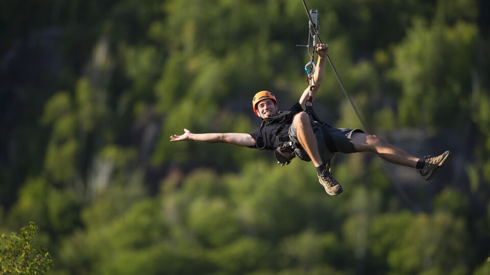 Un homme vêtu de noir avec un casque orange survole la cime des arbres à l'aide d'une tyrolienne dans les Laurentides.