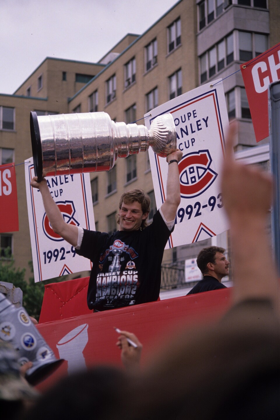 Un homme tient un trophée à bout de bras devant une foule et sourit.