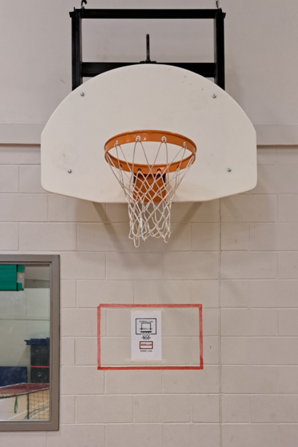 Un panier de basketball dans un gymnase au-dessous duquel on voit un rectangle rouge avec une affiche d'un autre panier