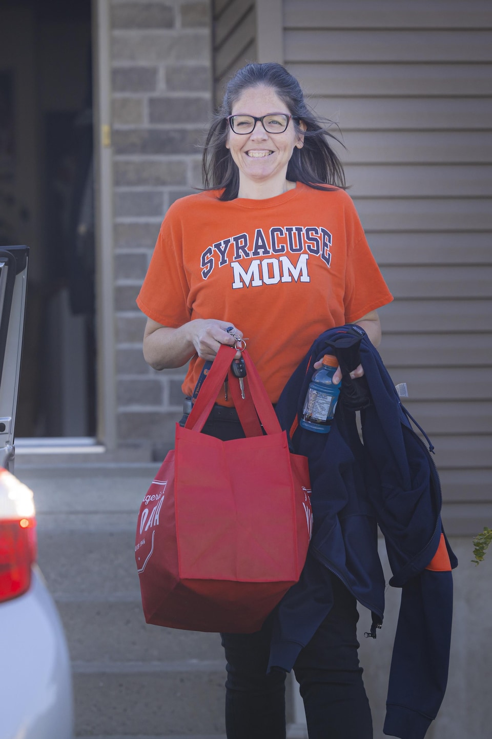 Une femme avec un chandail orange va déposer un sac dans le coffre de sa voiture