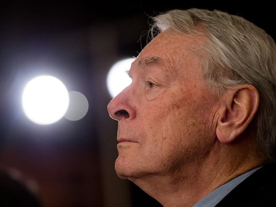 A man in close-up profile is looking straight ahead, seriously, with lights in the background