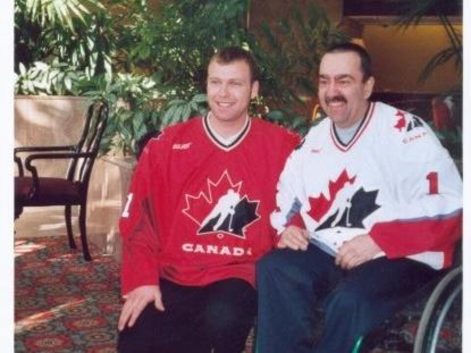 Un homme est accroupi à côté d'un autre homme assis dans un fauteuil roulant. Tous deux portent un chandail de l'équipe canadienne de hockey.