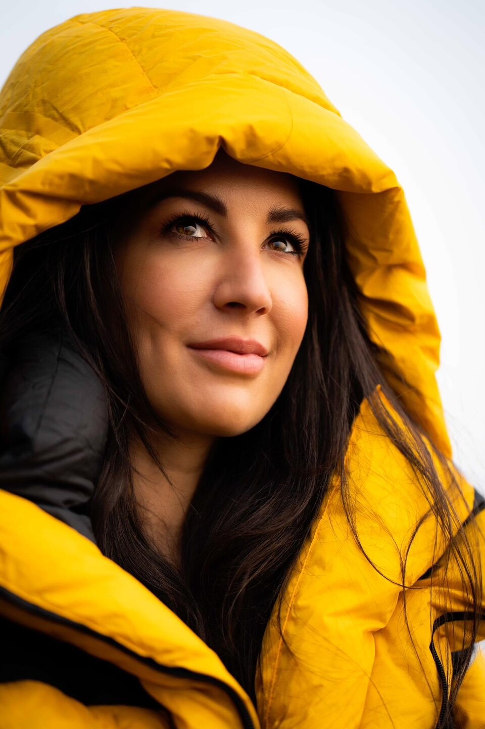 Mujer de cabello oscuro mira hacia adelante, vistiendo una gabardina con capucha amarilla. 