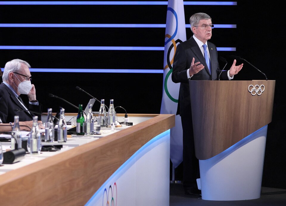 One man speaks with his hands open behind a lectern, the other is listening, sitting at a table.