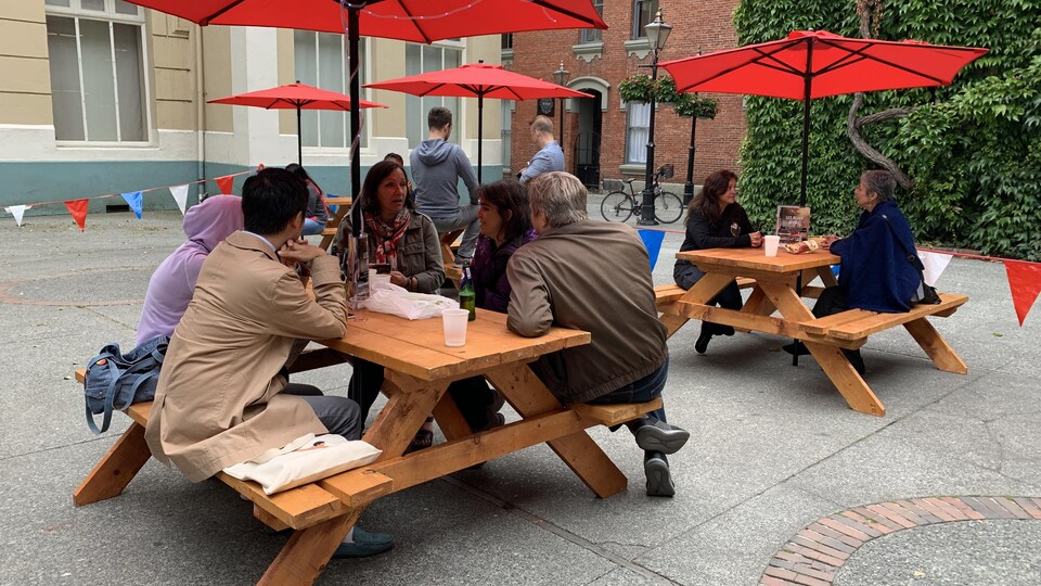 Des groupes d'amis sont installés à des tables en bois sous des parassols et boivent de la bière en discutant en français.