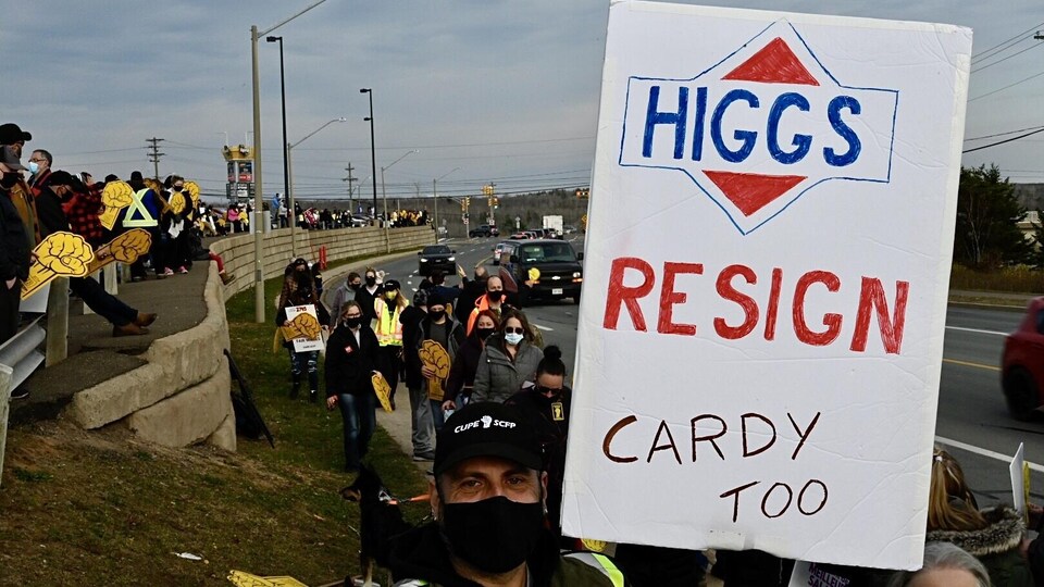 Manifestant tenant une affiche: Higgs Resign, Cardy Too.