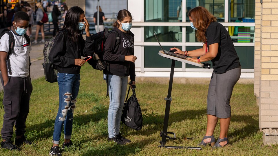 À l'extérieur de l'école, des élèves s'adressent à un membre du personnel qui consulte un ordinateur.