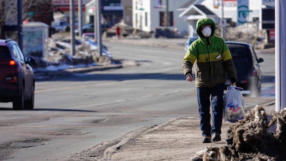 Un homme avec un masque sur le nez et la bouche marche dans une rue sale en transportant plusieurs sacs de plastique.