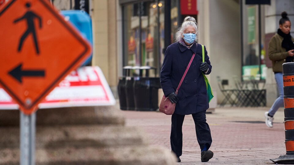 Une femme portant le masque chirurgical marche au centre-ville; une autre piétonne, derrière elle ne porte pas le masque.