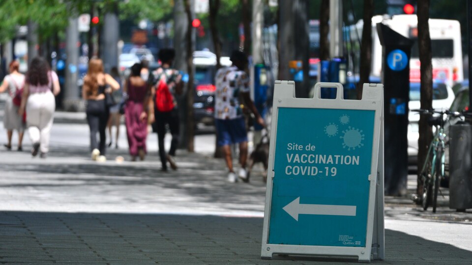 Des personnes marchant dans la rue. Au premier plan, un panneau indique la direction d'un centre de vaccination.