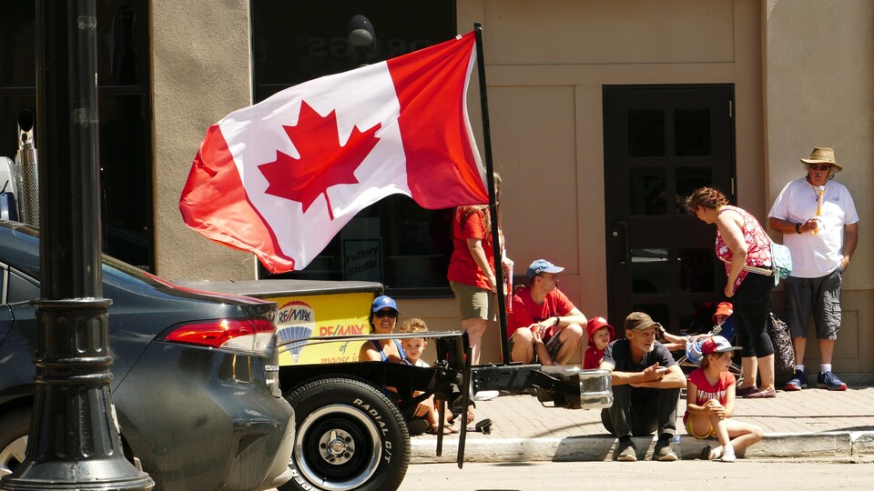 Des citoyens avec un grand drapeau du Canada regardent un défilé de voitures.