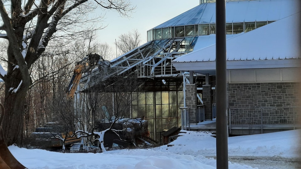 Une pelle mécanique procède à la démolition d'un bâtiment vitré en hiver.