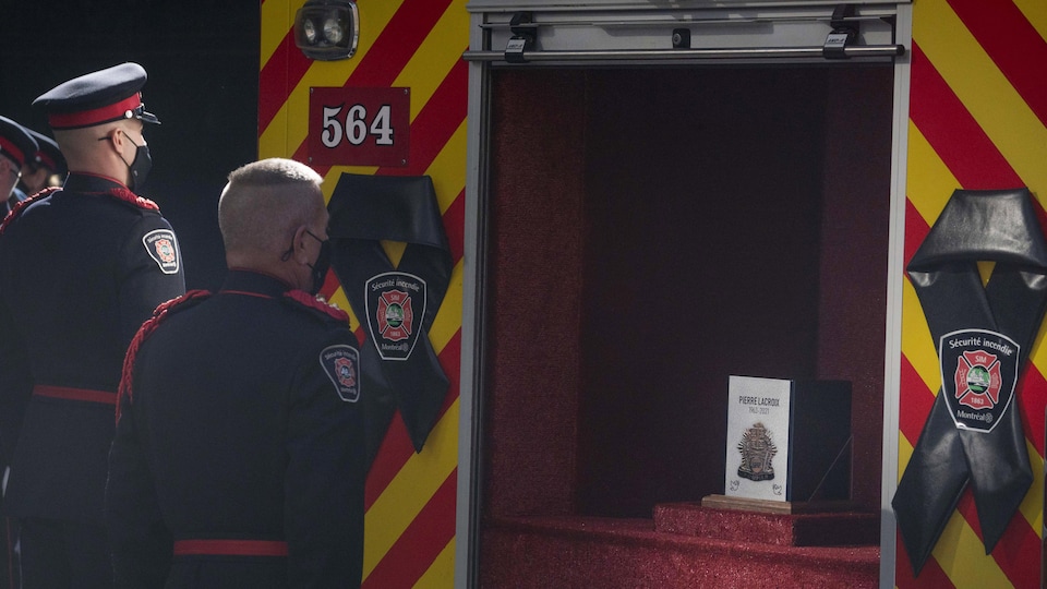Deux pompiers se recueillent devant une urne disposée de manière à rappeler un camion de pompiers.
