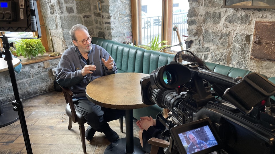Paul Piché est assis à une table, et il discute. 