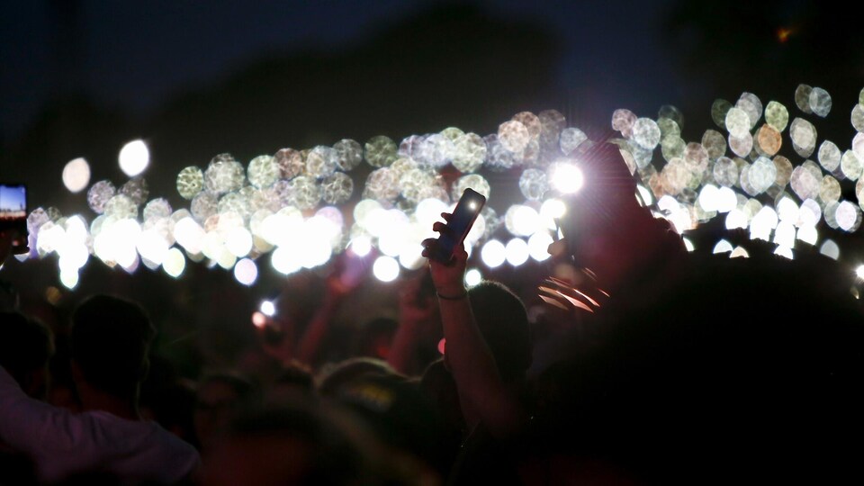 Une foule de spectateurs dans le soir, avec des lumières, et leur téléphone dans leur main