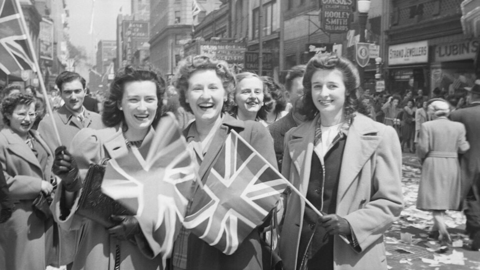 Trois jeunes femmes dans la foule qui célèbre le jour de la Victoire dans la rue Sainte-Catherine, à Montréal. Cette manifestation populaire souligne la capitulation allemande et la fin imminente de la Seconde Guerre mondiale.