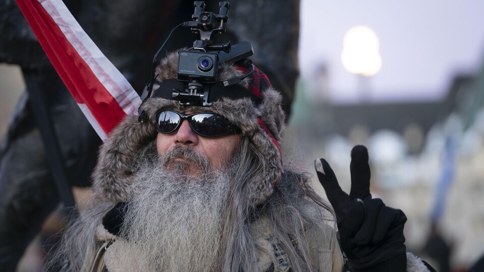 Un homme à la longue barbe fait un signe de paix de la main gauche.