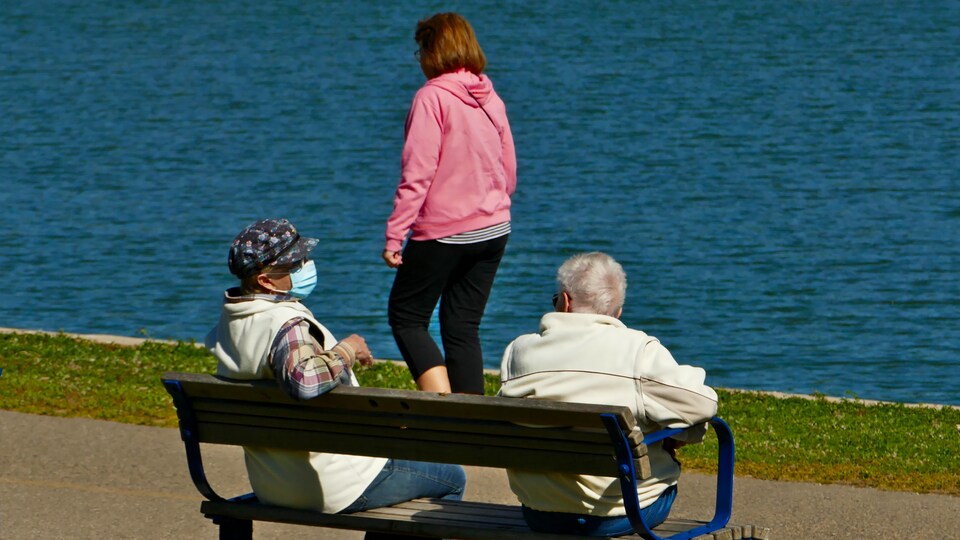 Des personnes en bordure d'un plan d'eau, dont deux assises sur un banc qui portent un masque.