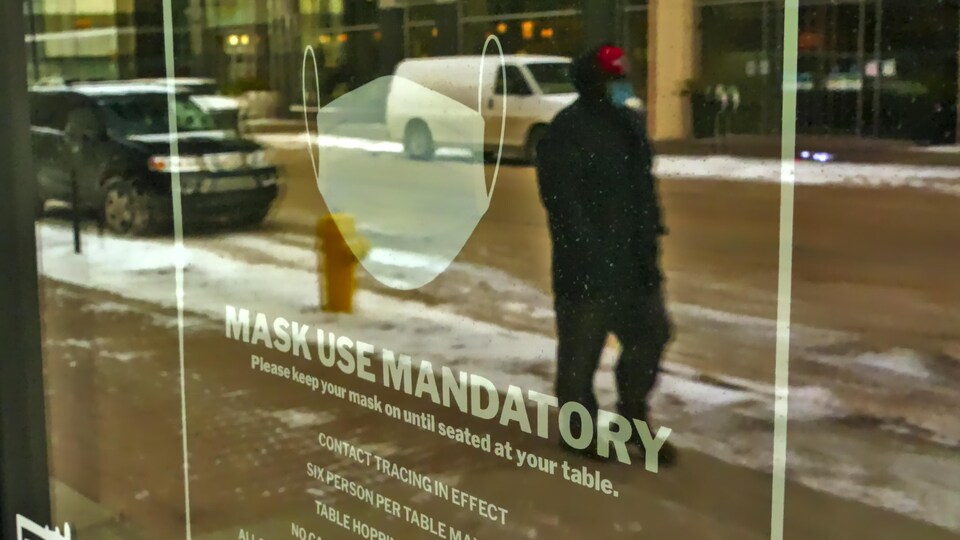 Le reflet d'un homme portant un couvre-visage dans une fenêtre dans laquelle on peut y lire des consignes sur le port du masque. 