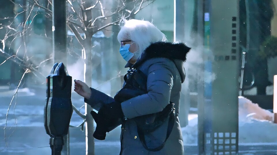 Une femme masquée, dans les rues de Regina, s'apprête à enfoncer une pièce dans un parcomètre.