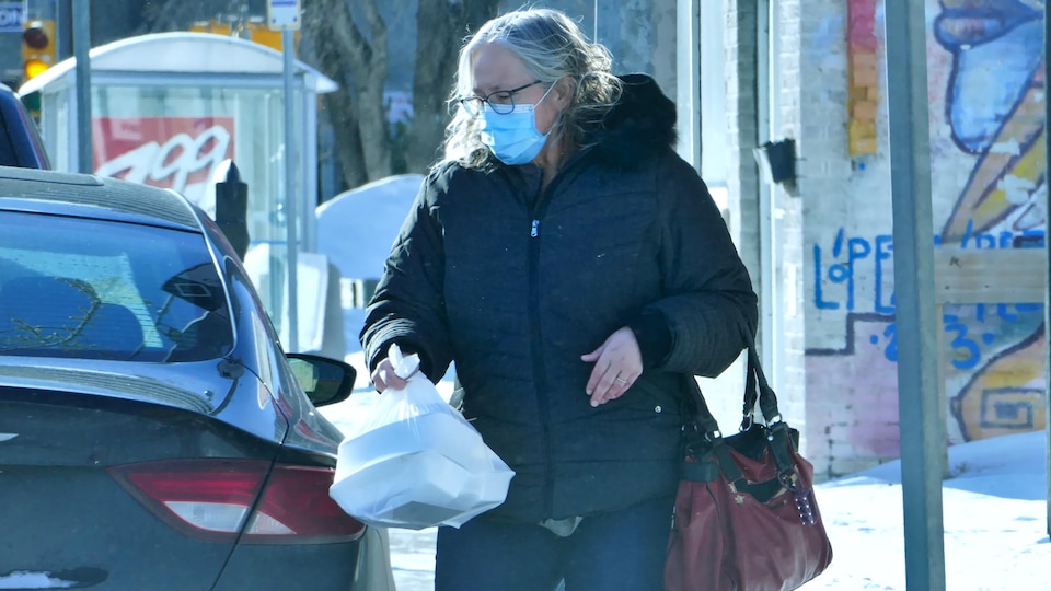 Une dame avec un masque porte des courses à sa voiture dans la rue.