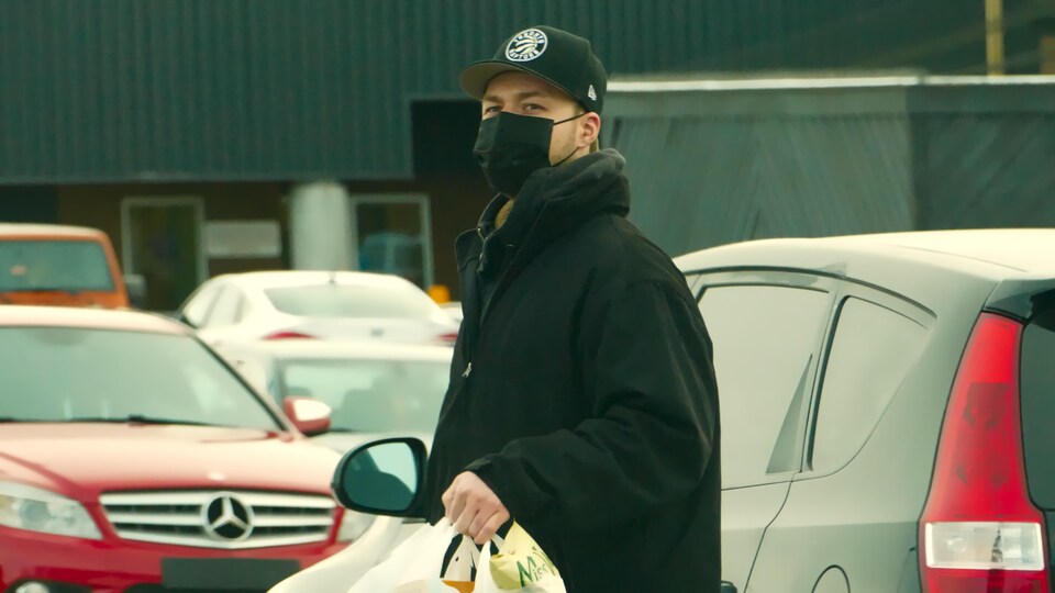 Un homme marche dans la rue avec un masque et une casquette. Il porte des sacs plastiques de course avec lui.
