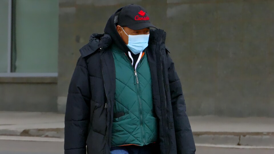 Un homme marche dans la rue avec un masque sur le visage et une casquette.