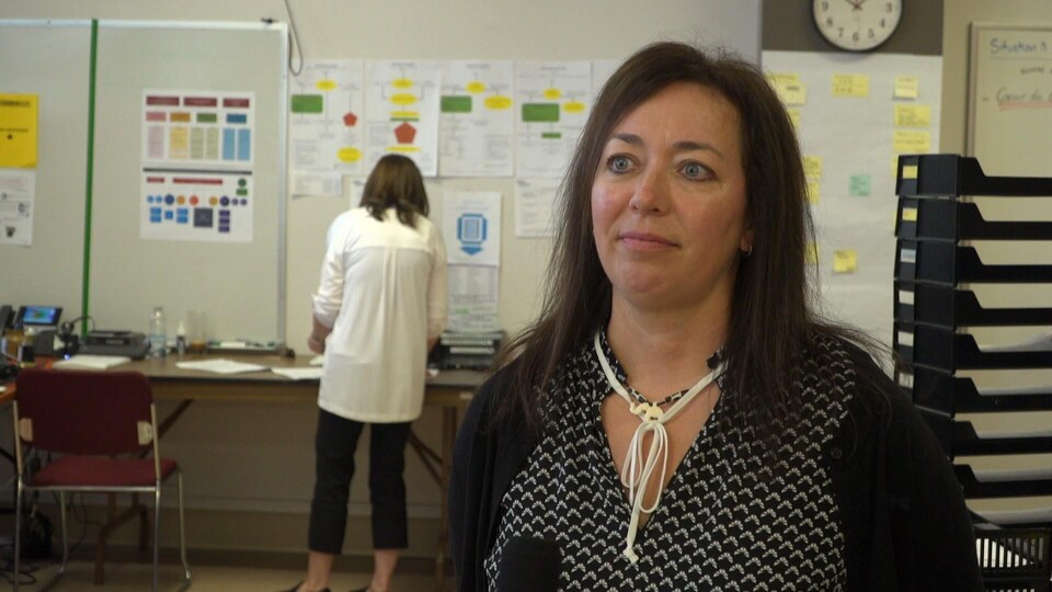 Isabelle Beaulieu accorde une entrevue dans un local. À l’arrière-plan, une femme, photographiée de dos, manipule des dossiers devant une table collée contre un mur.