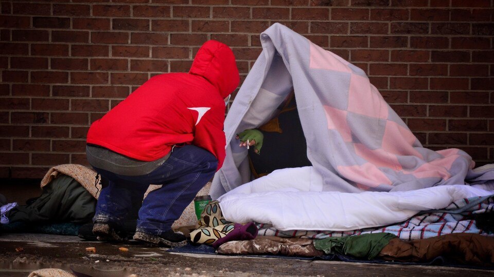 Une personne en-dessous de couvertures sur le trottoir, qui parle avec une autre personne de dos.