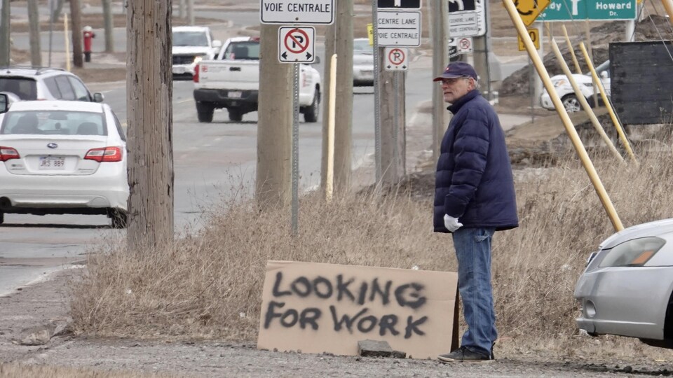 Un homme debout le long de la rue à côté d'un panneau improvisé sur lequel il est écrit : Je cherche un emploi.