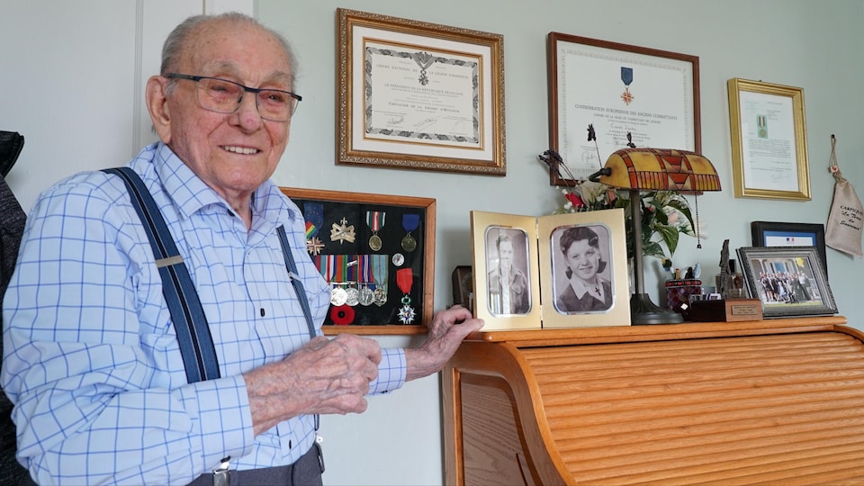 Homme âgé qui pose devant des médailles, des photos et certificats encadrés, dont un qui provient de l'Ordre national de la légion d'honneur.