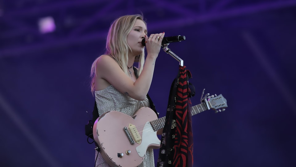 The singer puts her right hand on her microphone while singing.  She carries her guitar over her shoulder. 